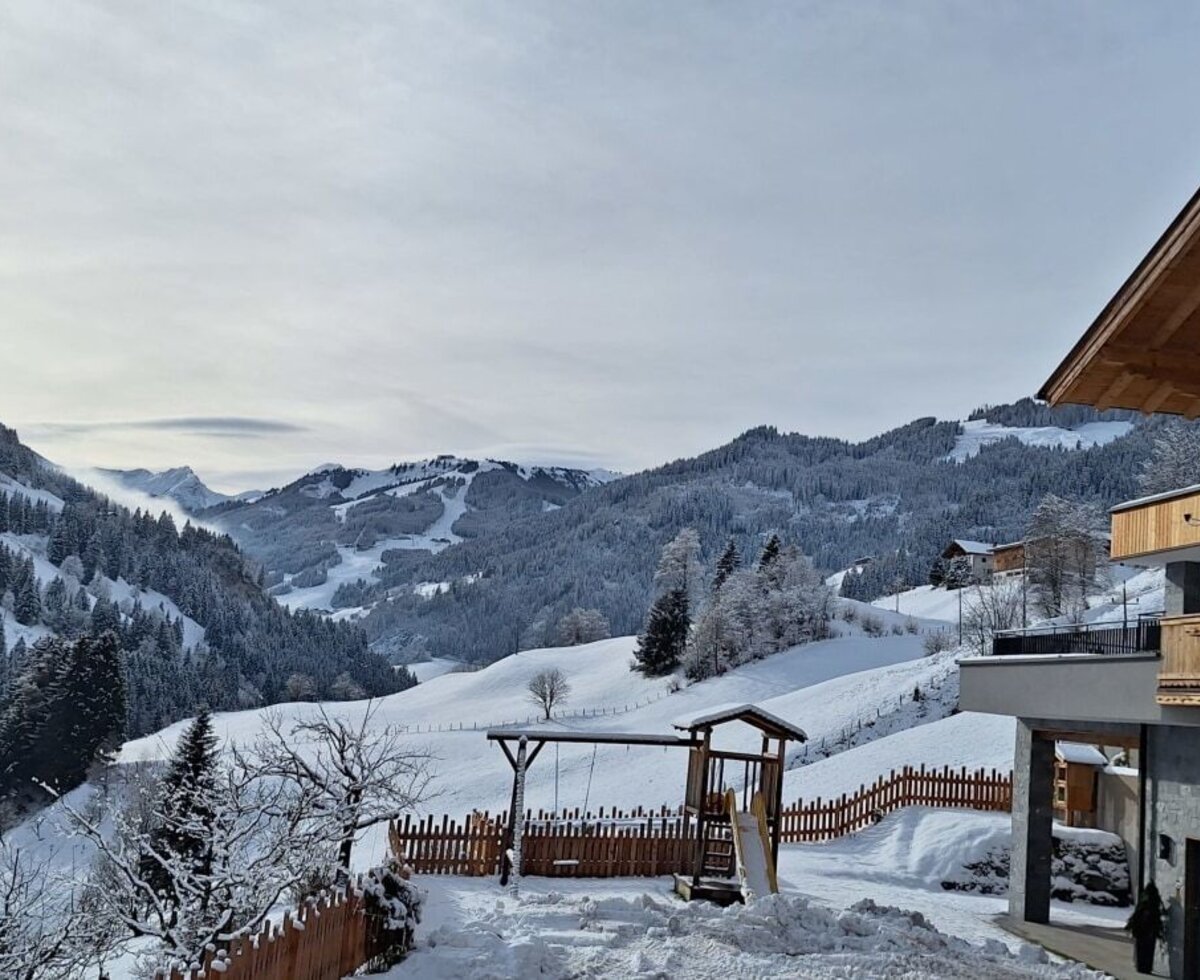 The exterior of the farm house featuring wooden balconies, a snow-covered children's playground, and views of the surrounding snowy mountains.