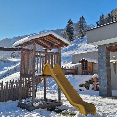 The Bauernhof's children's playground with a swing and slide, surrounded by snowy hills, and a covered outdoor seating area.
