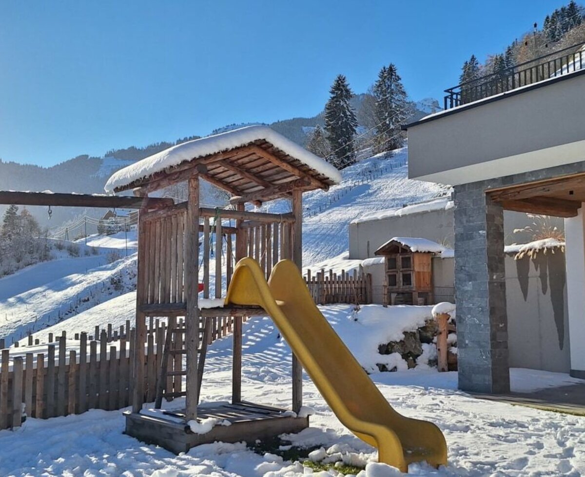 The Bauernhof's children's playground with a swing and slide, surrounded by snowy hills, and a covered outdoor seating area.