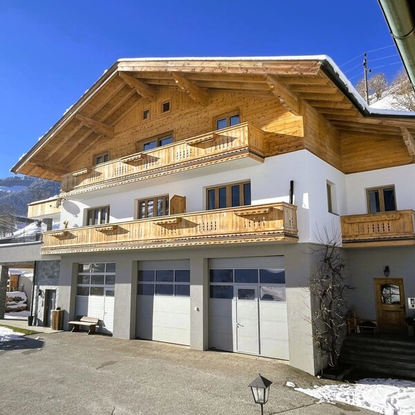 Exterior view of the Bauernhof with wooden balconies, a white facade, and garage doors, surrounded by snow.