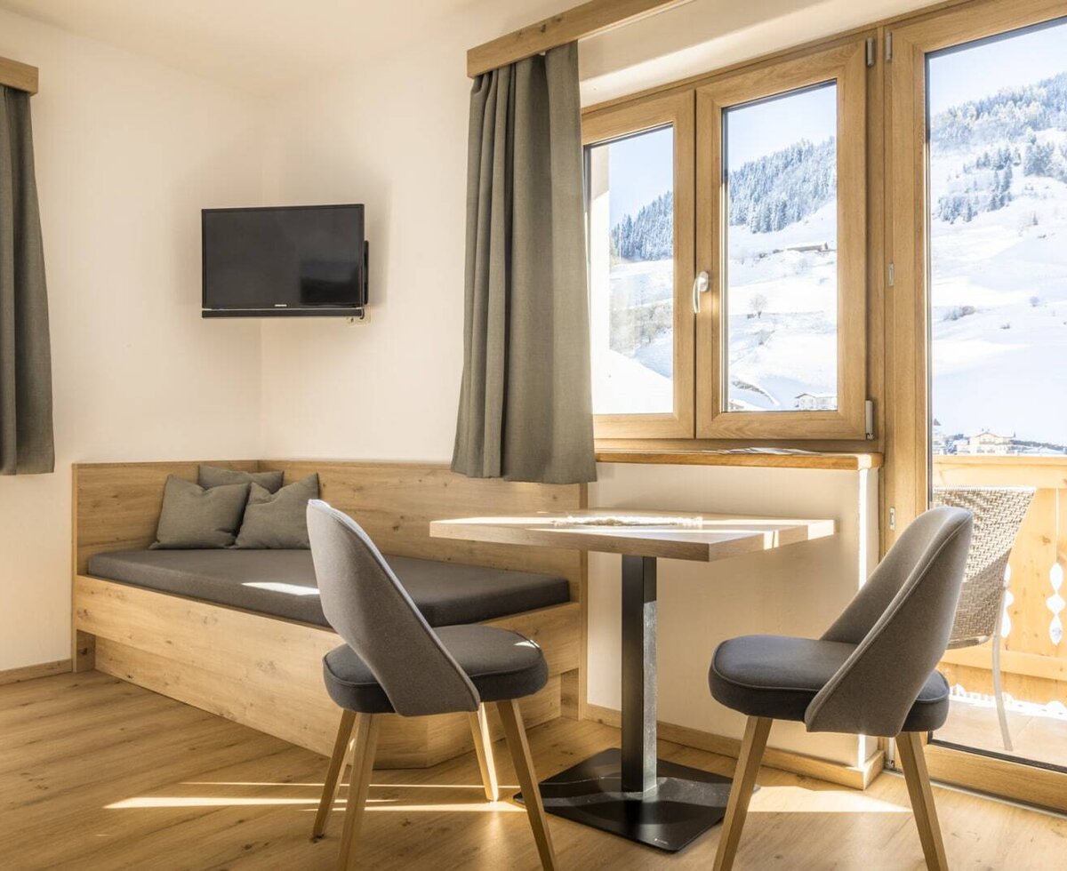 Room with a wooden bench, a dining table for two, a wall-mounted TV, and balcony access offering a view of the snowy landscape.