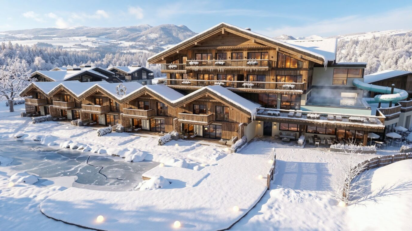 Exterior view of the Bauernhof in winter, featuring multiple buildings, a heated outdoor pool, a water slide, and a frozen pond.
