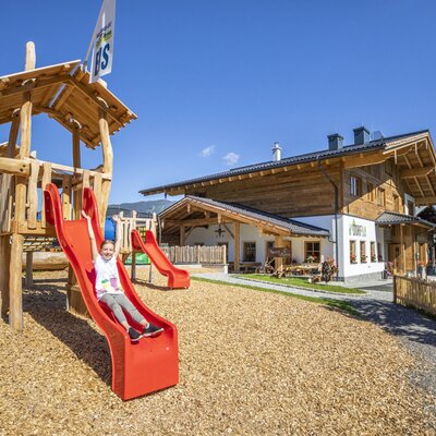 The farmhouse features a children's playground with a wooden climbing structure, red slides, and a wood chip base.