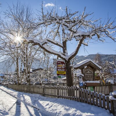 The snow-covered exterior of the farm house, featuring a path, wooden fence, and signs for the property and a milk vending machine, against a mountain backdrop.