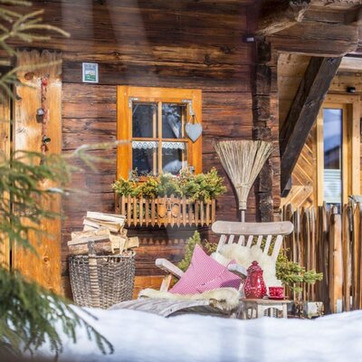 Wooden exterior of the farm house featuring a decorated window, a basket of firewood, a lounge chair with a sheepskin rug, and an outdoor sauna access.