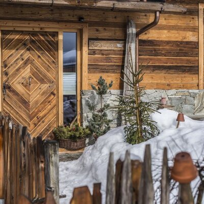 The exterior of the farm house, constructed from wood, includes a sauna entrance and another patterned wooden door, surrounded by snow.