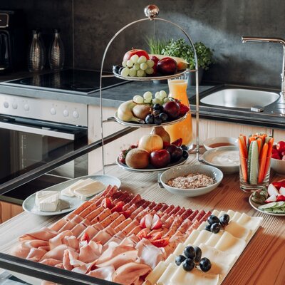 The Farm House kitchen features a stove, oven, sink, and coffee machine, alongside a breakfast spread with cold cuts, cheeses, fresh fruit, and vegetables.