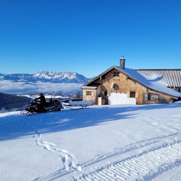 The snow-covered Schoberhütte farmhouse in winter, featuring a snowmobile and views of the Untersberg mountains.