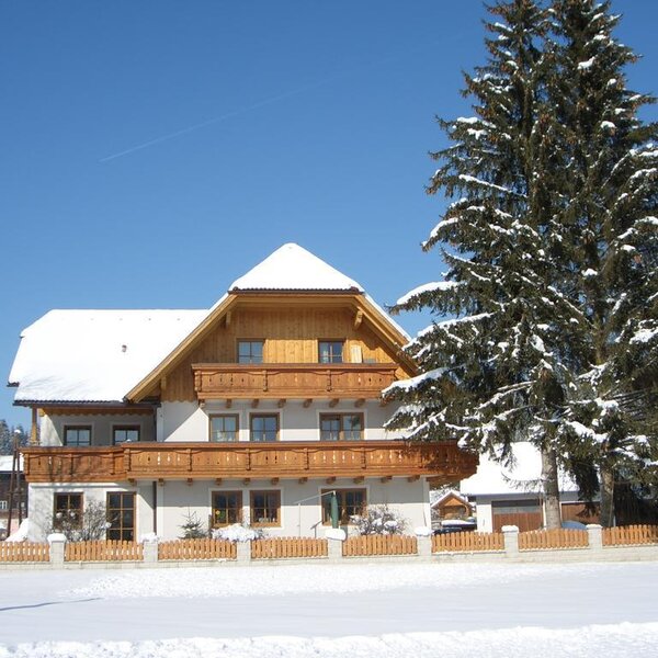 The Farm House exterior in winter, featuring wooden balconies, a snow-covered roof, and a large fir tree in front.