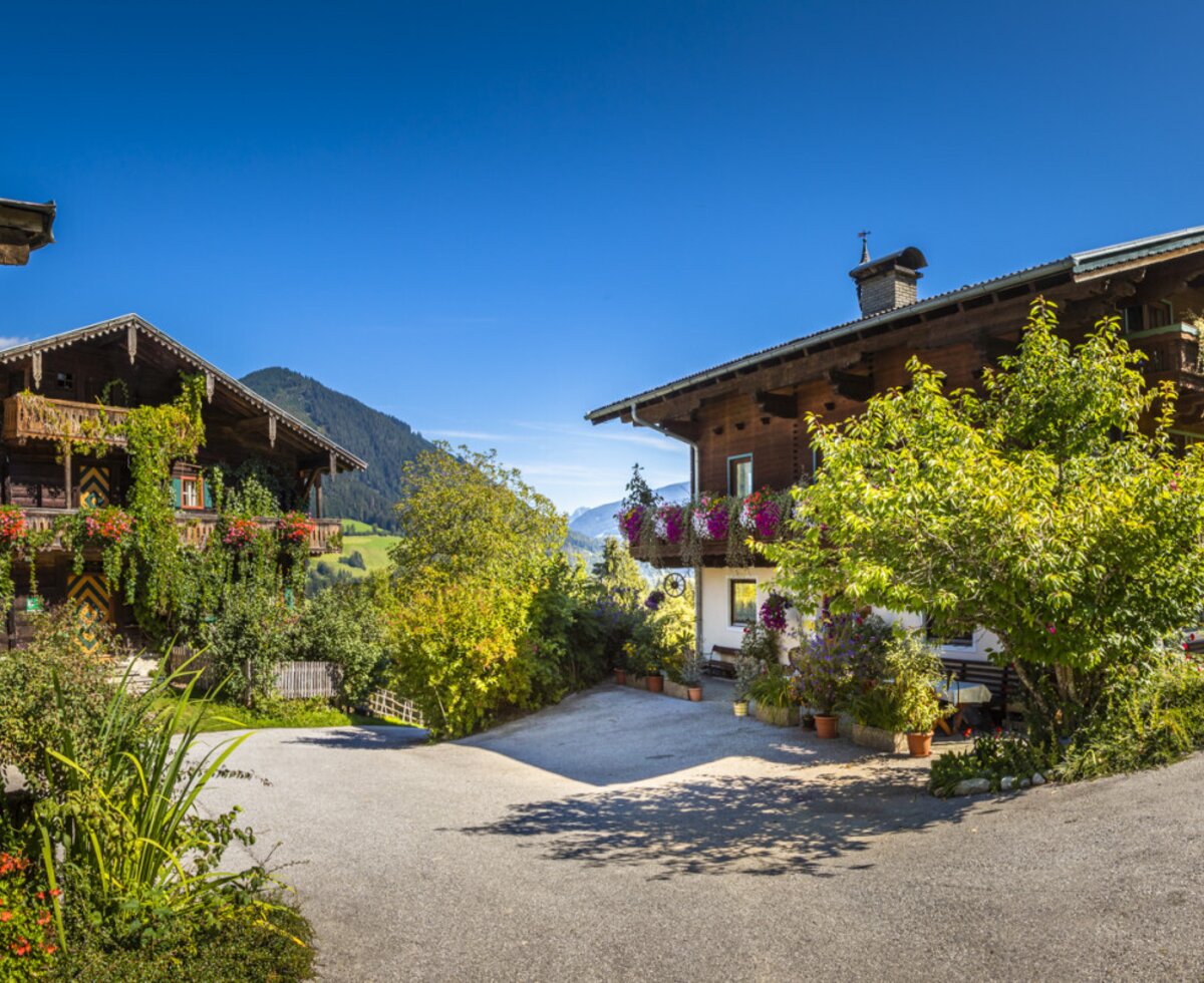 The farmhouse exterior, showcasing two traditional buildings with ornate wooden balconies and colorful flower boxes, set against a backdrop of green mountains.