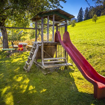 A children's playground at the farmhouse featuring a wooden climbing frame, a red slide, and a swing set.