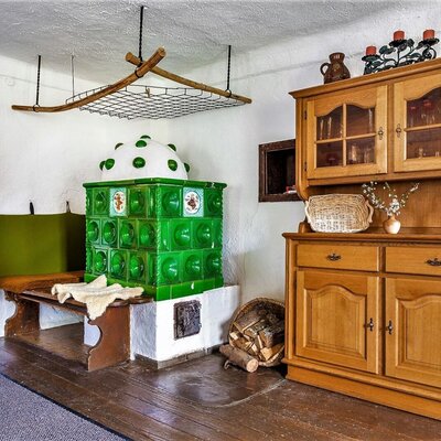 The farmhouse features a traditional green tiled stove with an adjacent wooden bench for guests, and a classic wooden hutch.