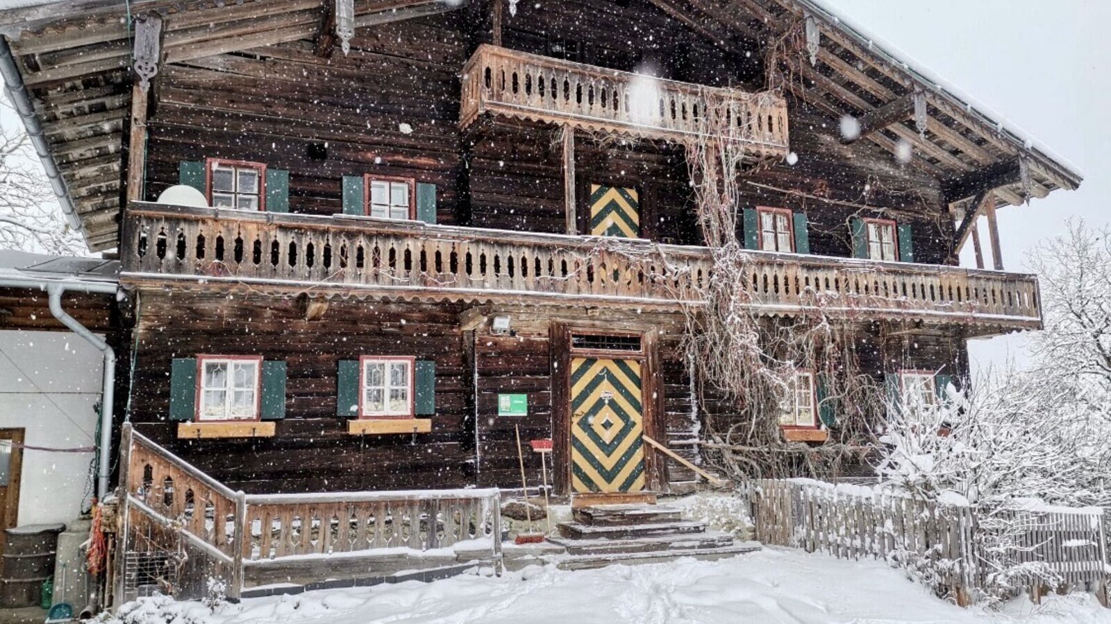 The wooden farmhouse exterior, featuring balconies with ornate railings, green-shuttered windows, and a distinctive patterned main door, pictured in the snow.
