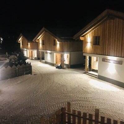 The illuminated wooden exterior of the farm house at night, featuring multiple units and a snow-covered path.