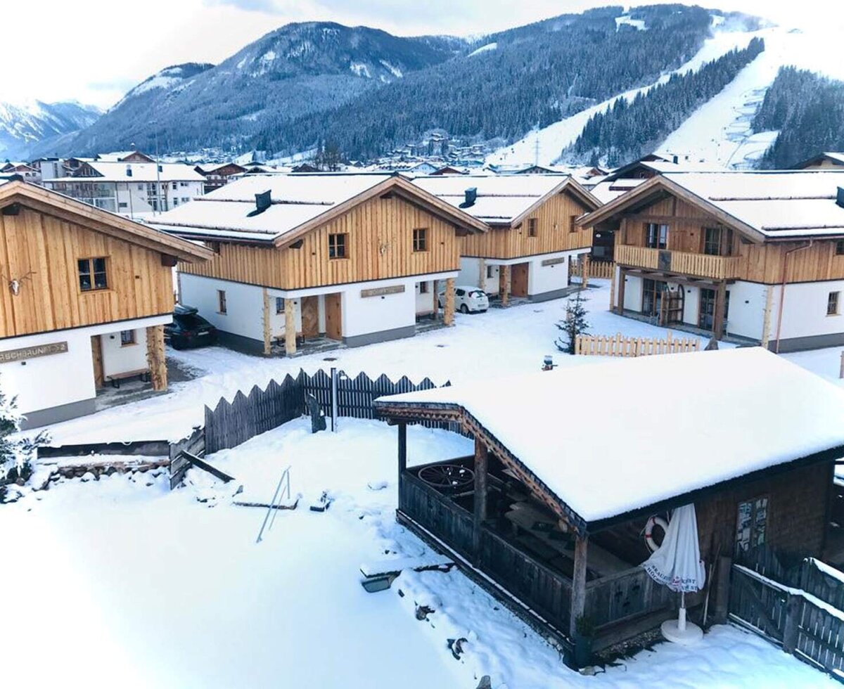 A group of farmhouses with snow-covered roofs, set against a backdrop of mountains and ski slopes.