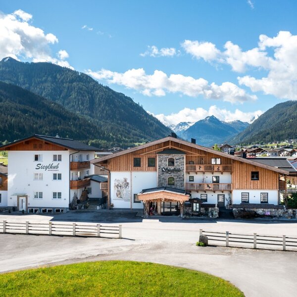 The Siegthub Farm House exterior, featuring traditional architecture with wooden elements and balconies, set against a backdrop of green mountains.
