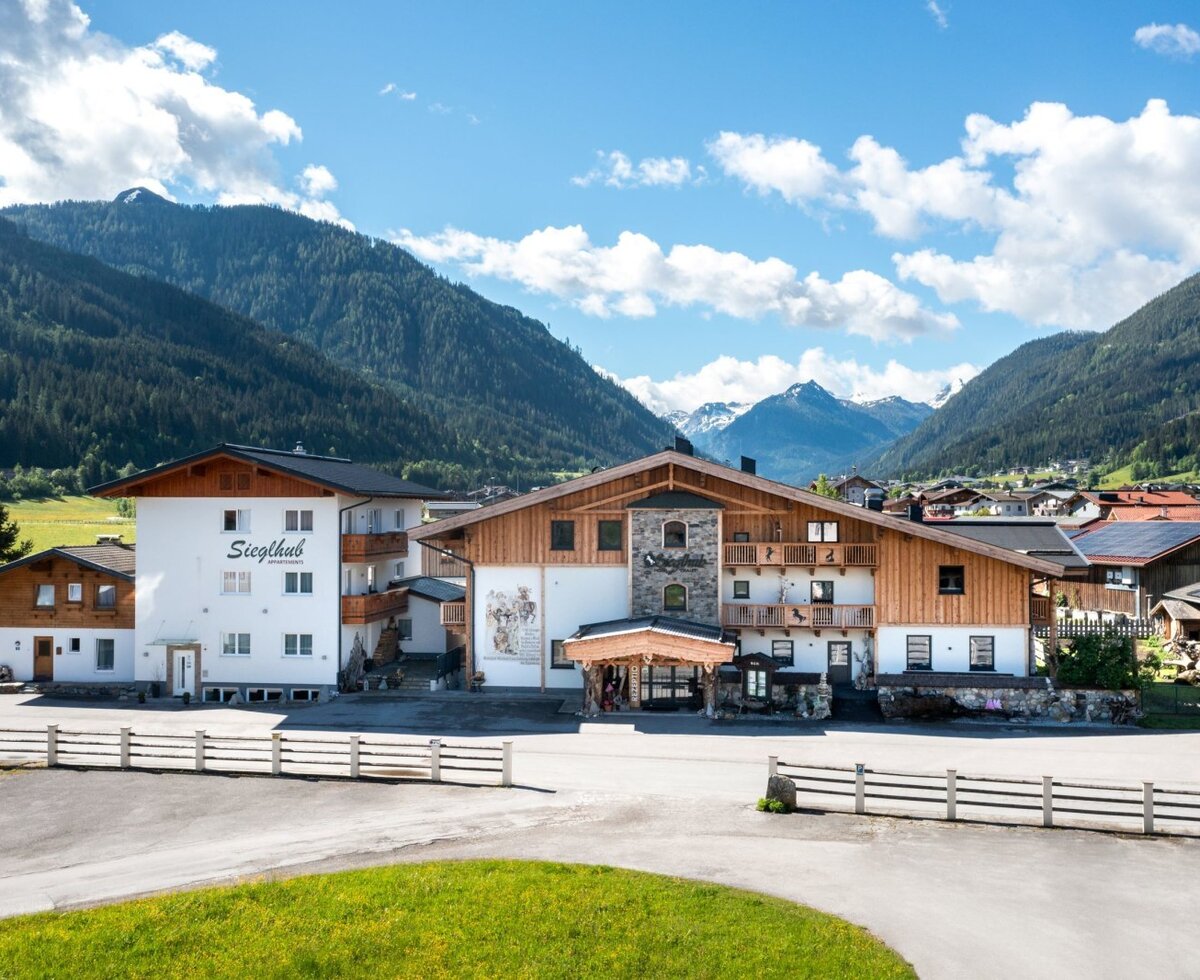 The Siegthub Farm House exterior, featuring traditional architecture with wooden elements and balconies, set against a backdrop of green mountains.