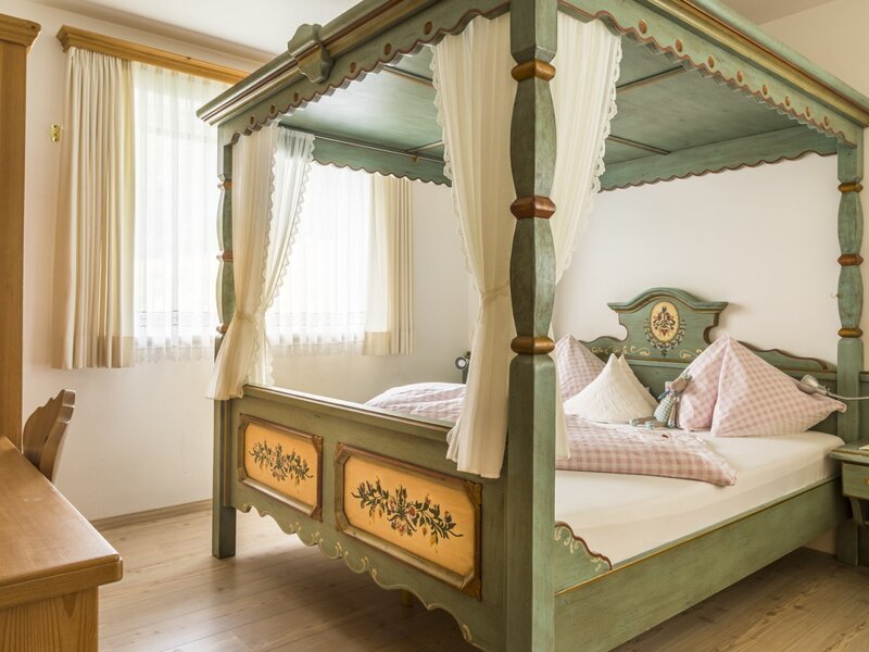 Bedroom in the Farm House with a green four-poster bed featuring decorative painted details and drapes, a wooden desk, and a window.