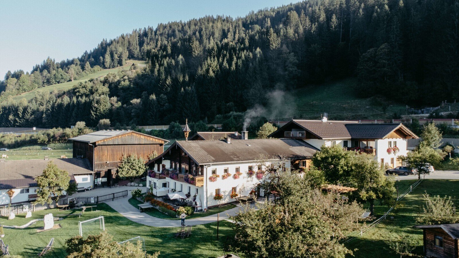 The farmhouse complex features a main building with balconies and flower boxes, a large wooden barn, and a spacious lawn with a children's play area, all set against a forested hillside.