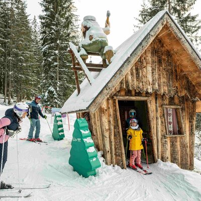 A family skiing on the slope next to a wooden hut, surrounded by snow-covered trees.