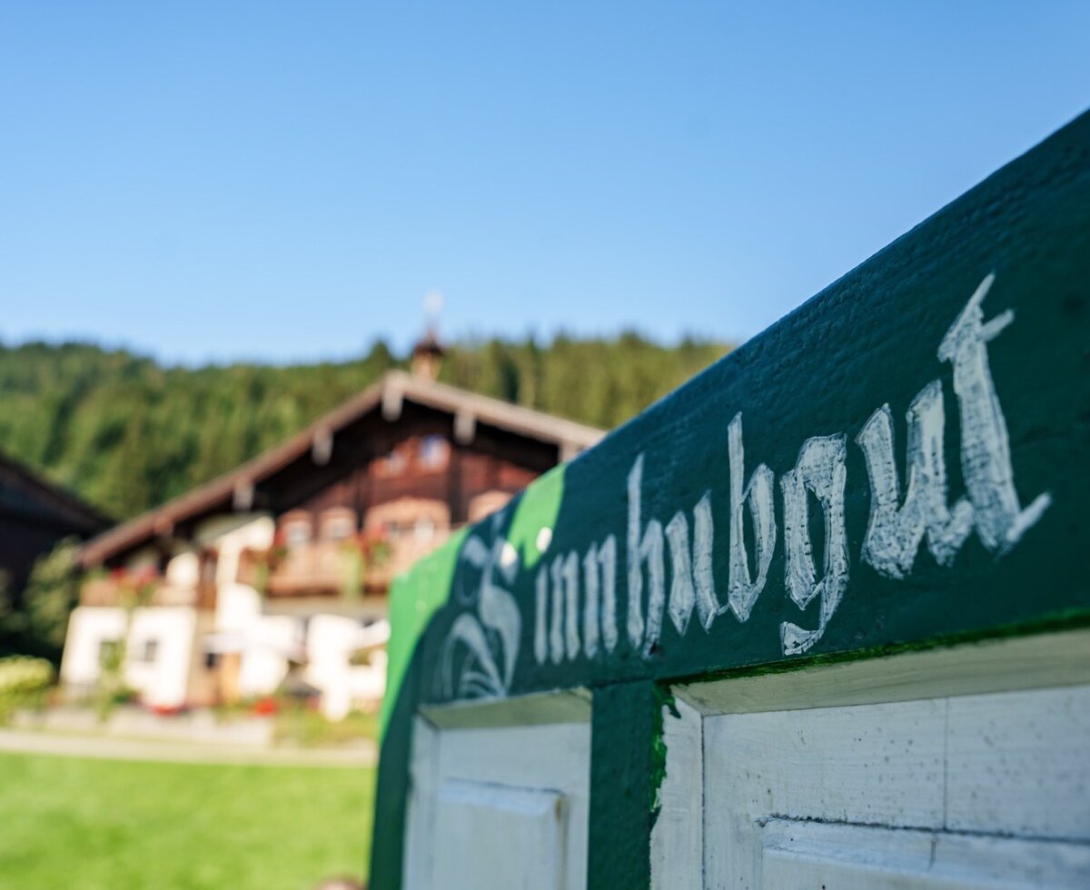 Sign for the 'Sinnhubgut' farmhouse with the main building and wooded hills in the background.