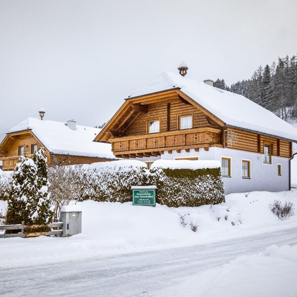 The Farm House exterior during winter, featuring a snow-covered roof, wooden balcony, and surrounding landscape.