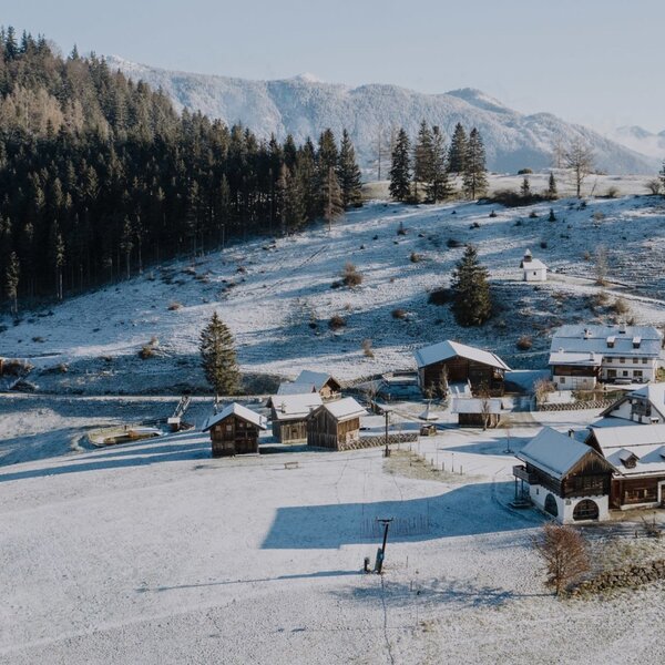 The farmhouse in a snow-covered alpine landscape, surrounded by traditional buildings, forests, mountains, and a nearby small ski lift.