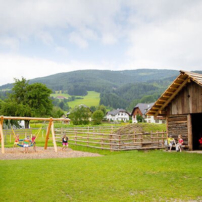 Children's playground at the farmhouse with swings and children playing, next to a wooden barn and mountain scenery.