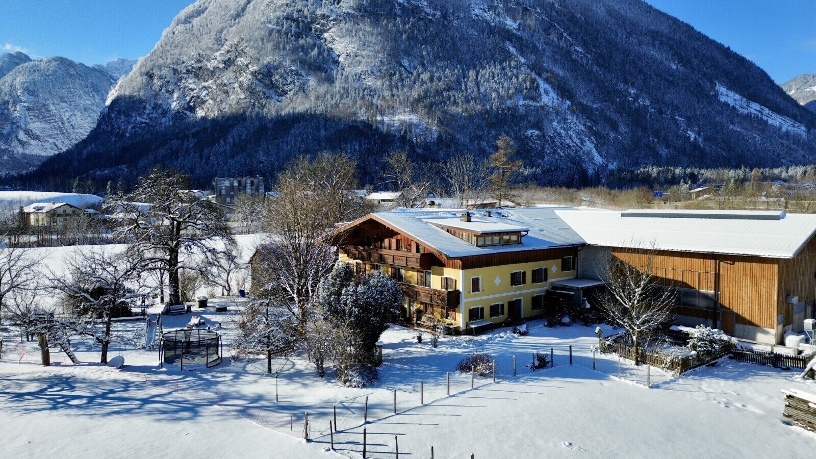The farmhouse in winter, featuring snow-covered roofs, wooden balconies, and a yard with playground equipment, surrounded by mountains.