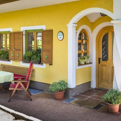 The yellow exterior of the Farm House features an arched entrance, windows with wooden shutters and flower boxes, and an outdoor patio with a table, chairs, and a wooden bench.