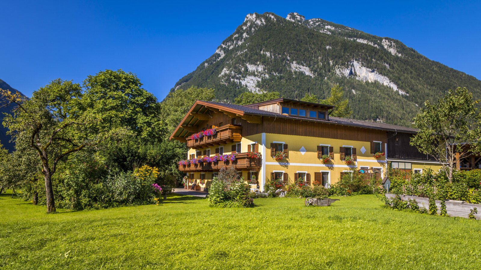 The Farm House exterior with a yellow facade, wooden balconies adorned with flowers, a green lawn, and mountains in the background.