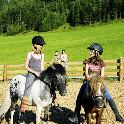 Children wearing helmets ride ponies in a fenced area at the farm house, with green hills and a forest in the background.