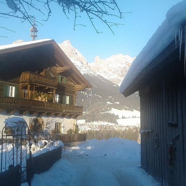 The snow-covered Farm House exterior with traditional wooden balconies, an adjacent outbuilding, and a view of distant snowy mountains.