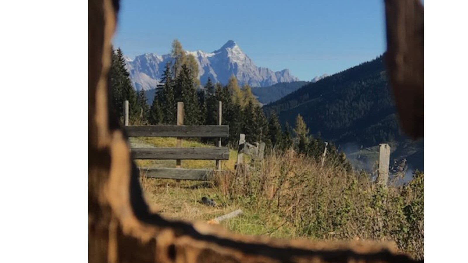 Mountain view, framed by wood, with trees and a fence in the foreground.