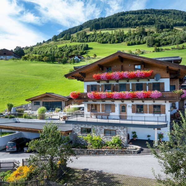 Exterior view of the farmhouse featuring flower-decorated balconies, a carport, and a green hillside.