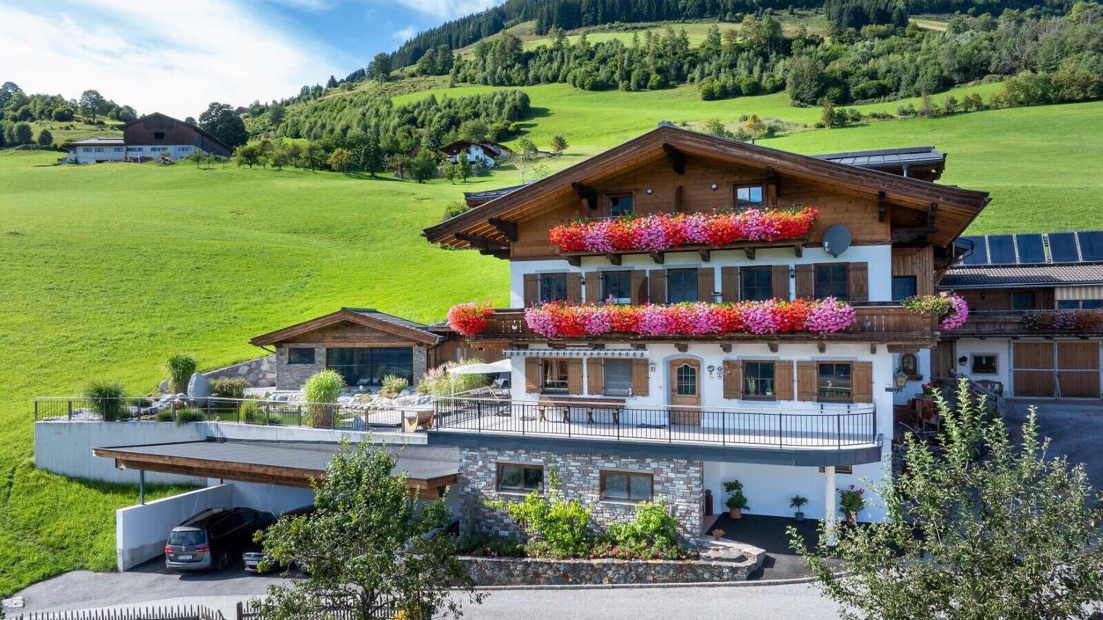 Exterior view of the farmhouse featuring flower-decorated balconies, a carport, and a green hillside.