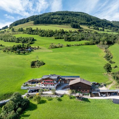 The farmhouse complex, surrounded by green meadows and forested mountains, featuring multiple buildings and solar panels.