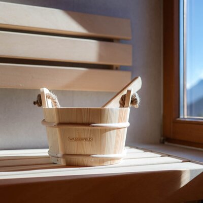 Wooden sauna bucket and ladle in the farmhouse's sauna, next to a window with a mountain view.
