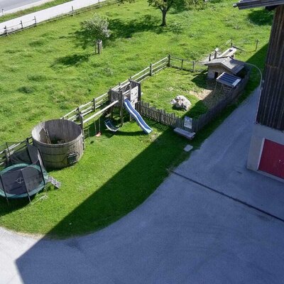 The farm house features a children's play area with a slide, swings, trampoline, and a large wooden barrel, located next to the building with red garage doors.