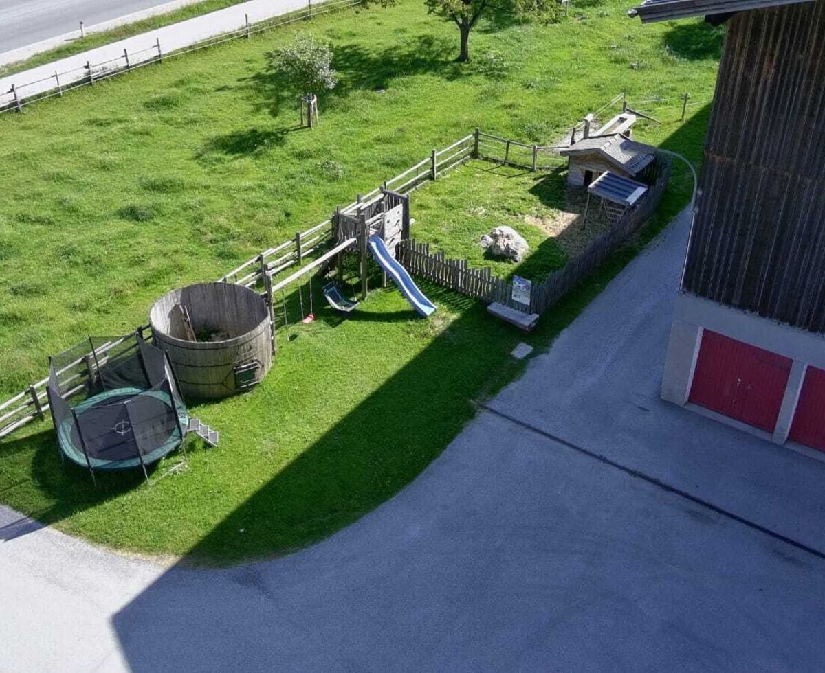 The farm house features a children's play area with a slide, swings, trampoline, and a large wooden barrel, located next to the building with red garage doors.