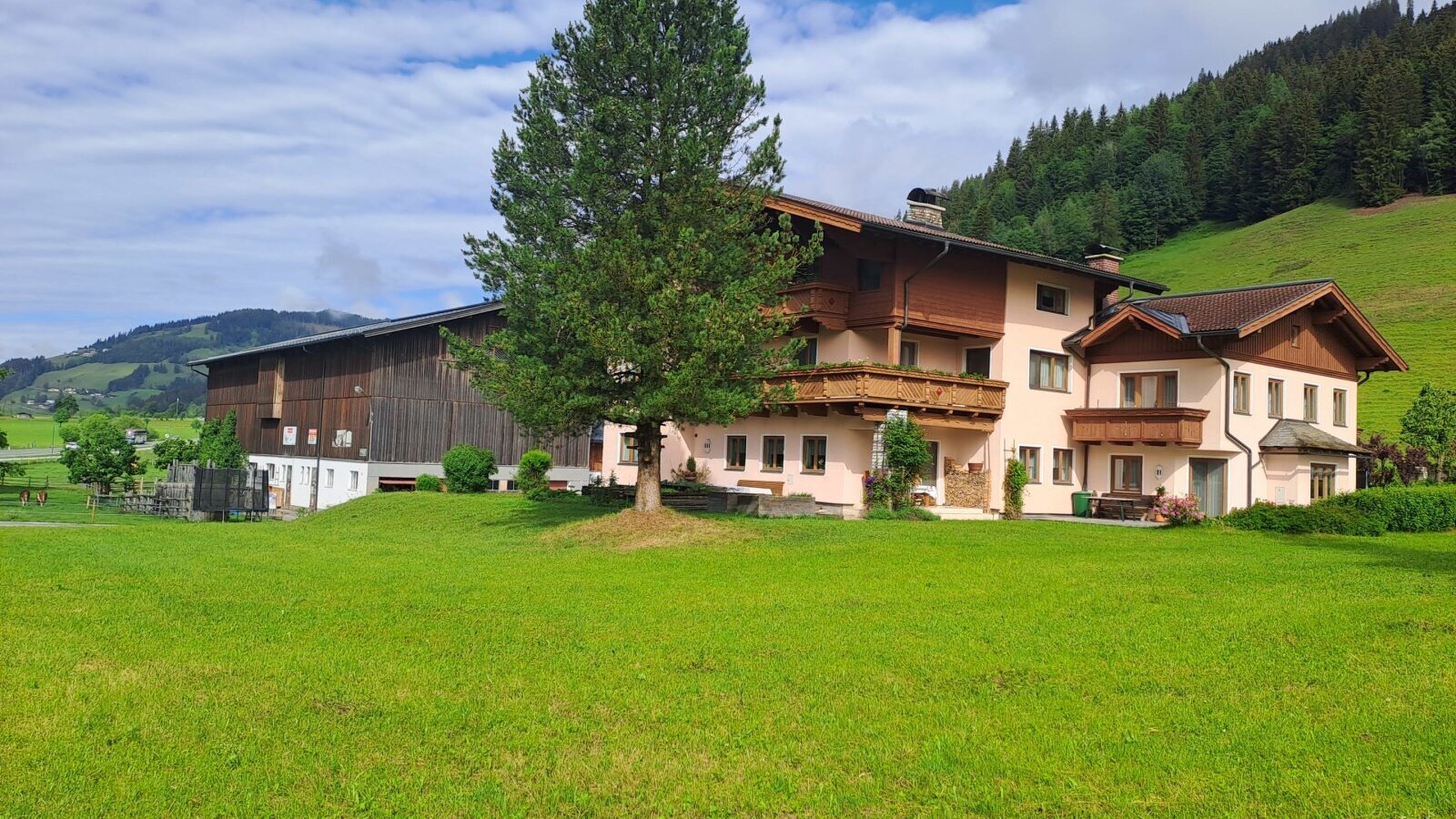 Exterior view of the farmhouse with balconies, a large lawn, and an adjacent barn against a mountain backdrop.