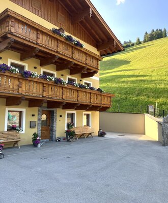 The farm house features a yellow exterior, multiple wooden balconies adorned with flower boxes, and a paved courtyard with benches, set against a green hillside.