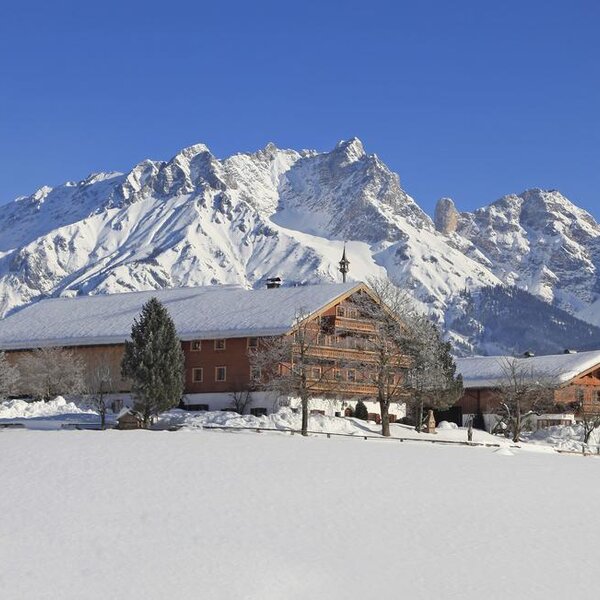 The traditional wooden Farm House stands in a snow-covered landscape with prominent snow-capped mountains in the background.
