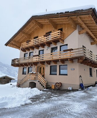The renovated farmhouse exterior, featuring wooden balconies and an entrance staircase, surrounded by snow.
