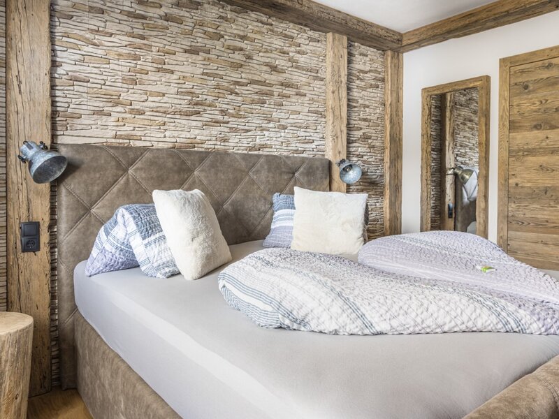 Bedroom of the Ferienwohnung with a double bed, padded headboard, stone wall, wooden beams, and a mirror.