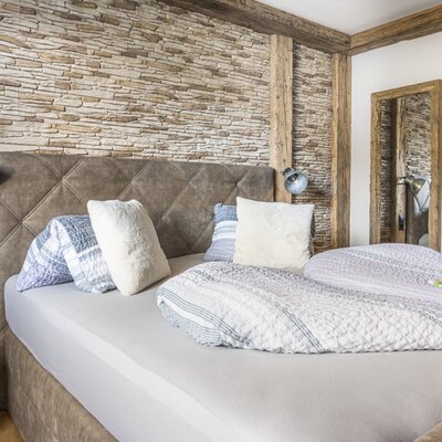 Bedroom of the Ferienwohnung with a double bed, padded headboard, stone wall, wooden beams, and a mirror.