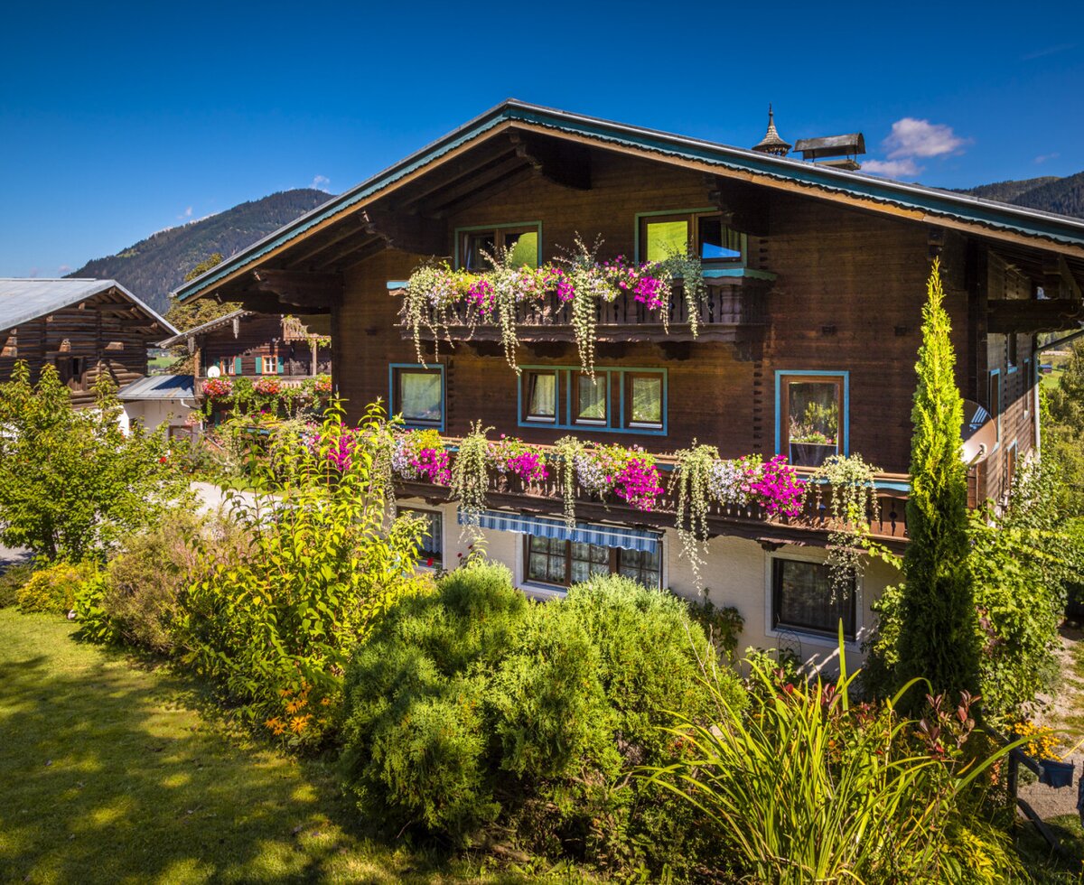 The Farm House exterior displays traditional wooden architecture, multiple balconies with flower boxes, and a surrounding garden against a mountain backdrop.