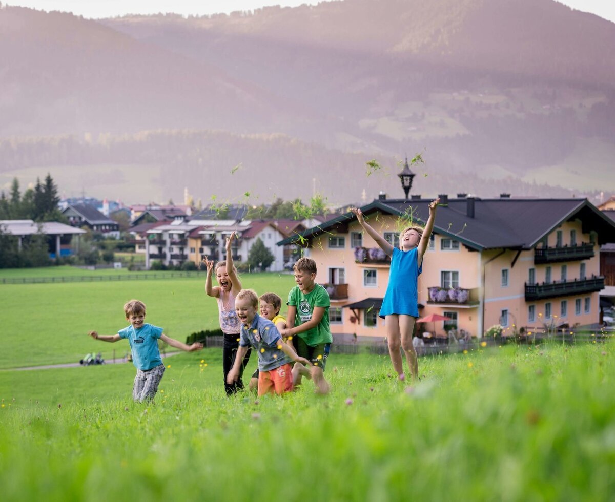 Children playing in the spacious outdoor fields of the Farm House, set against a backdrop of mountains and a village.
