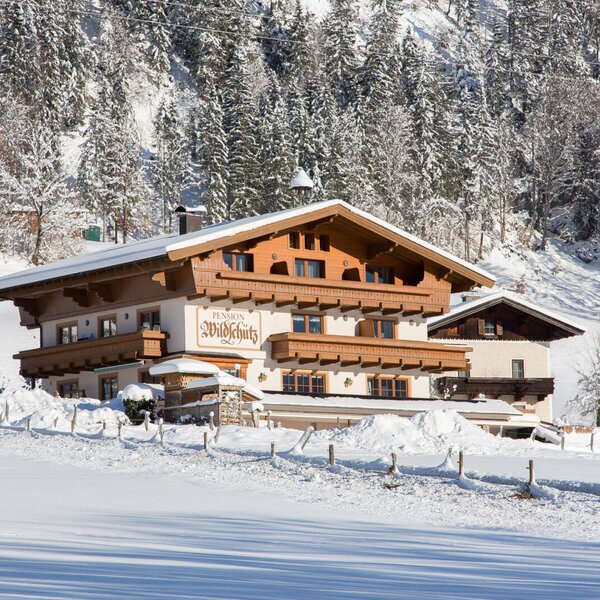 The farm house 'Pension Wildschütz' exterior in winter, surrounded by snow-covered fields and a forested mountain.