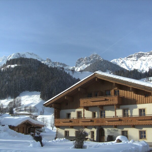 The traditional farm house exterior in winter, featuring snow on its balconies and roof, set against a backdrop of snow-covered mountains and forests.
